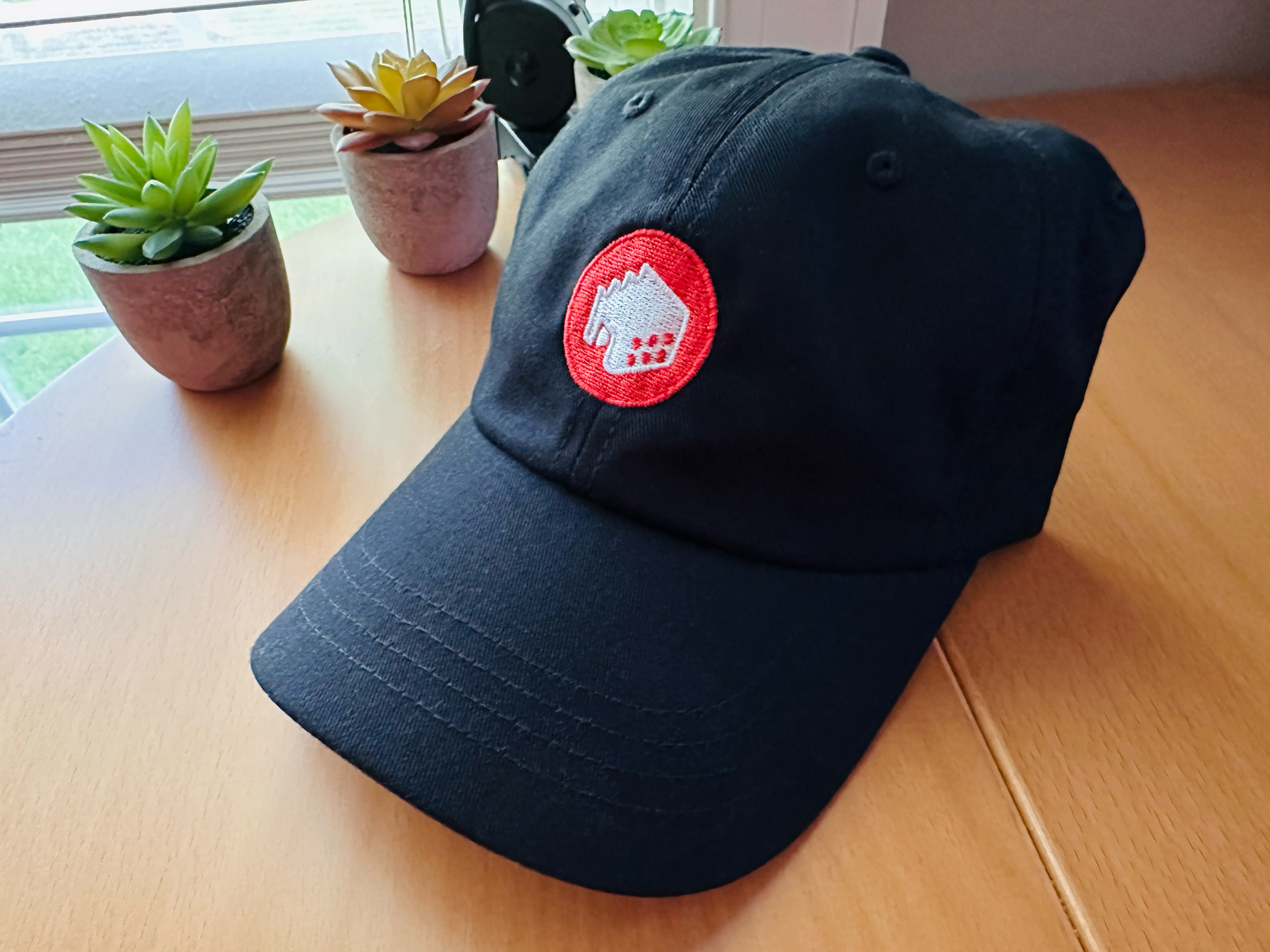 A black baseball cap sitting on a wood desk embroidered with the red and white logo of The Iconfactory.